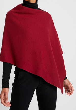 Femme portant un poncho asymétrique en laine rouge sur un col roulé noir et un pantalon noir, debout devant un fond clair uni.