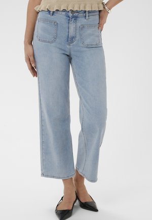 Kvinde iført lyseblå cropped vidde jeans med forlommer, kombineret med sorte spidse flade sko og en beige top.
