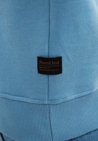 Sudadera azul claro con ribetes en el dobladillo y los puños, con una etiqueta de tela negra que dice "Petrol Ind" en letras naranjas.