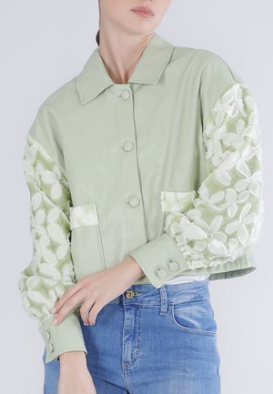 Giacca corta verde chiaro con bottoni frontali e maniche a sbuffo in pizzo floreale, abbinata a jeans blu su uno sfondo neutro.