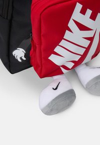 Rdeča in črna nahrbtnik z velikim belim logotipom "Nike", s plišastimi nogami in podrobnostjo risanih rok na strani.
