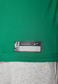 Camiseta deportiva verde hecha de una tela suave con una etiqueta de marca negra que presenta texto en blanco. Pantalones cortos grises con un detalle sutil del logo.