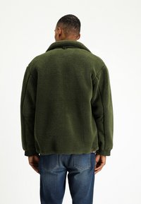 Zielona polarowa kurtka o teksturowanej powierzchni, w oversize'owym kroju, z zapięciem na guziki przy kołnierzu i elastycznymi mankietami, noszona z niebieskimi dżinsami.