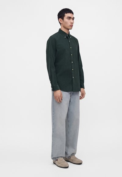 Jeune homme debout portant une chemise boutonnée vert foncé, un jean large bleu clair et des chaussures beiges à enfiler sur un fond blanc uni.