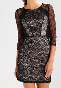 Černé krajkové šaty s dlouhými průhlednými rukávy, zdobené geometrickým vzorem a přiléhavým střihem. Texturované detaily po celé látce.