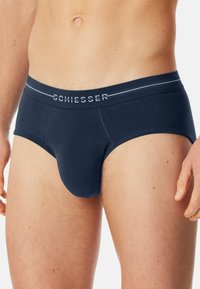 Mannelijk bovenlijf draag een marineblauwe Schiesser onderbroek met een gemerkte tailleband, zichtbaar abdomen en gedeeltelijk zichtbare handen tegen een effen achtergrond.