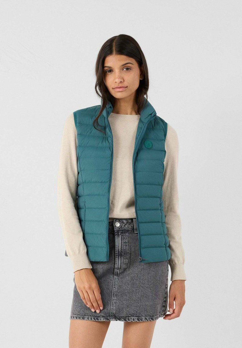 Gilet imbottito teal con colletto alto e zip frontale, indossato sopra un maglione crema. Il gilet presenta cuciture orizzontali e un dettaglio con il logo.