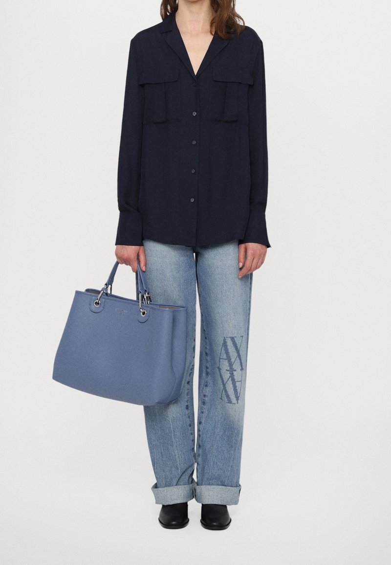 Personne portant une chemise noire à boutons, un jean bleu clair avec revers et logo « AX », des chaussures noires, tenant un grand sac à main bleu dans sa main gauche.
