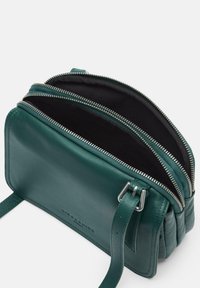 Zielona skórzana torba na ramię z trzema zamykanymi na zamek komorami, gładką fakturą, regulowanym paskiem i srebrnymi elementami metalowymi.