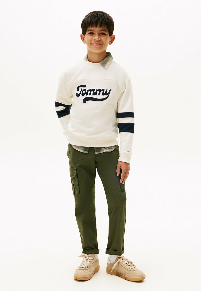 Witte sweatshirt met marineblauwe strepen, "Tommy" logo; olijfgroene cargobroek; beige sneakers met rubberen zolen; gelaagde kraag.