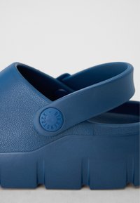 Blå träskor gjorda av texturerad plast med ett brett rem och en rund tå. Har en märkbar cirkulär logotyp och en tjock, räfflad sula.