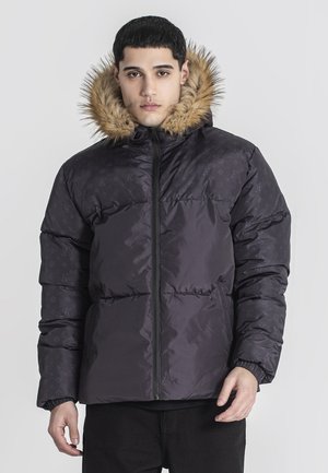 Junger Mann trägt eine schwarze Daunenjacke mit pelzgefütterter Kapuze und steht vor einem schlichten hellen Hintergrund.