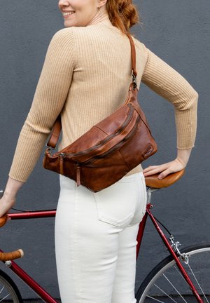 Pruun nahast õlakott mitme lukuga taskuga ja reguleeritava rihmaga, kantud üle risti, kombineeritud valgete pükstega ja beeži ribaga kampsuniga.