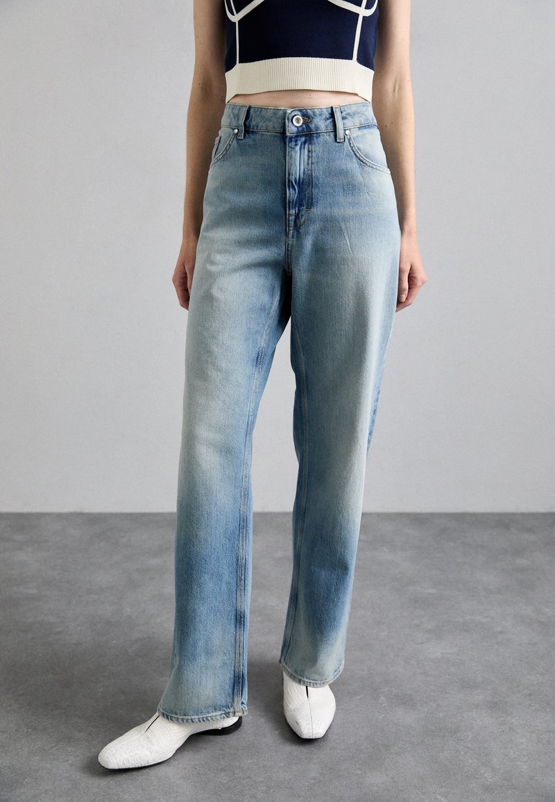 Jeans de mezclilla azul claro con un corte relajado, con cintura alta, un ligero desgastado y diseño clásico de cinco bolsillos. Combinados con zapatos blancos.