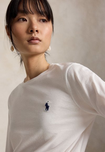 Νέα γυναίκα με σκούρα μαλλιά που φοράει λευκό πουκάμισο με μικρό σκούρο λογότυπο παίκτη πόλο, κοιτάζει ελαφρώς προς τα πλάγια σε απλό φόντο.