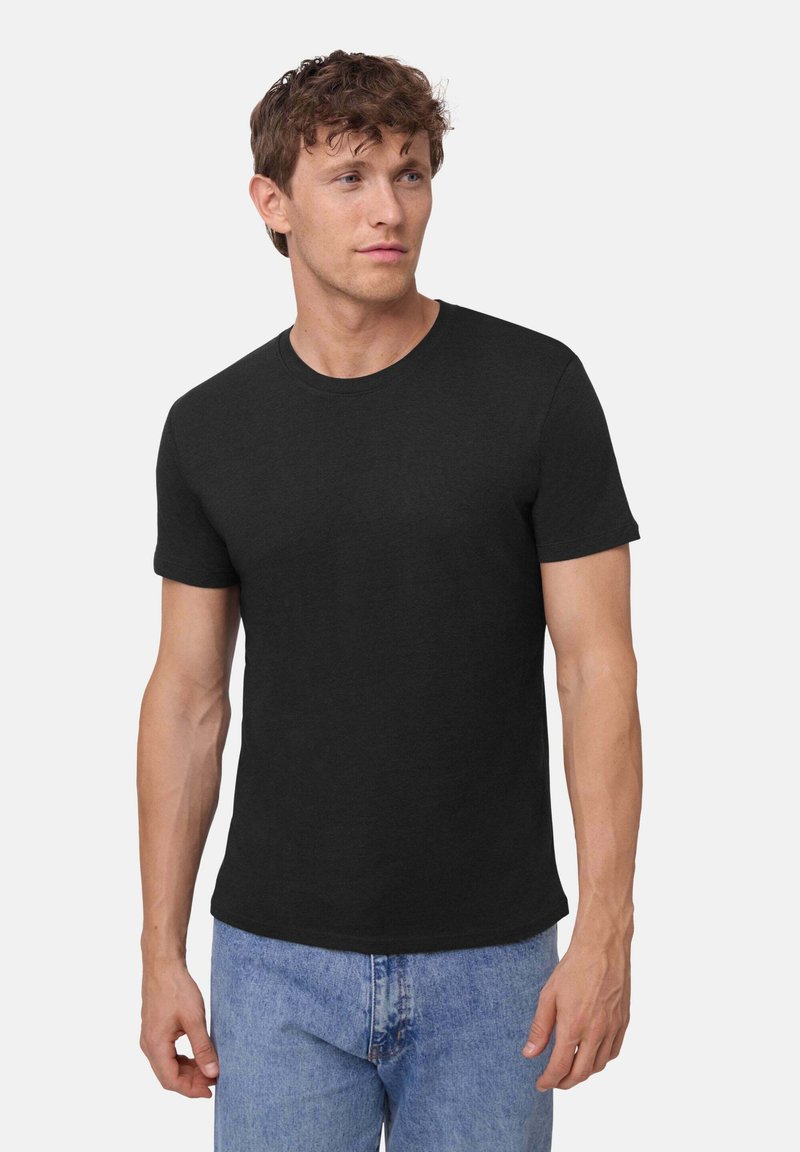 Man met kort krullend haar, gekleed in een nauwsluitend zwart T-shirt met korte mouwen en lichtblauwe jeans, kijkt iets naar links.