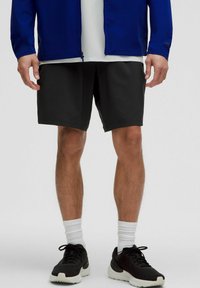 Man i svarta träningsshorts, vita crewstrumpor, svarta sneakers, en vit skjorta och en blå zip-up jacka som står framför en vit bakgrund.