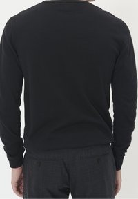 Mies, joka on pukeutunut mustaan pitkähihaiseen paita ja tummanharmaisiin ruutuhousuihin, kuvattu takaapäin yksinkertaista valkoista taustaa vasten.