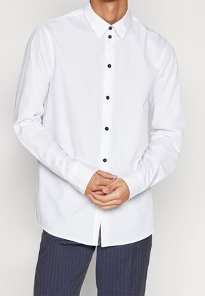 Homme portant une chemise blanche à manches longues avec des boutons noirs et un pantalon foncé à fines rayures, mains jointes devant.