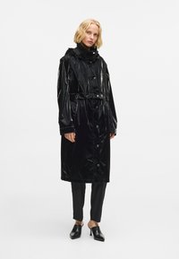 Melna gara lietus mētelis ar spīdīgu apdari, kuram ir sašauts jostasvietā, pogas, regulējama kapuce un garas piedurknes. Akcentēts ar melnām biksēm un apaviem.