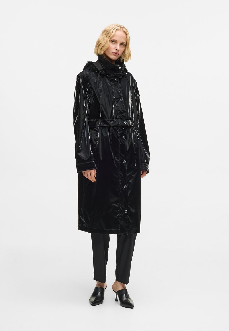 Melna gara lietus mētelis ar spīdīgu apdari, kuram ir sašauts jostasvietā, pogas, regulējama kapuce un garas piedurknes. Akcentēts ar melnām biksēm un apaviem.