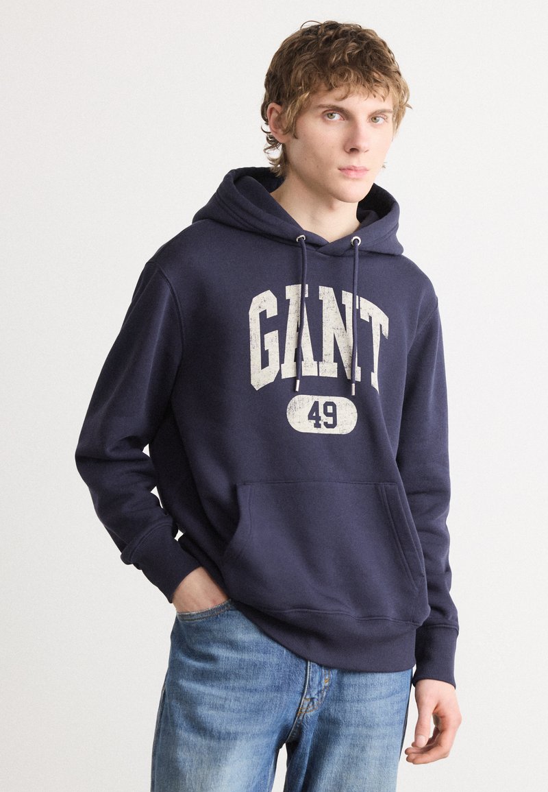 Mørkeblå hettegenser med frontlomme, snøring i hetta og hvit slitt logo tekst "GANT 49." Laget av myk stoff.