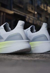 Paire de chaussures de course blanches Adidas UltraBoost avec des accents gris et des semelles épaisses et rembourrées sur une surface extérieure rugueuse.