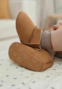 Brązowe zamszowe kapcie o miękkiej fakturze, z paskiem i wzorami w kształcie twarzy misia na podeszwie, umieszczone na jasnej, wzorzystej powierzchni.