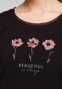 Camiseta negra con escote en forma de pico que presenta tres impresiones de flores rosas con centros negros, acompañadas del texto "HERMOZA como siempre" debajo.