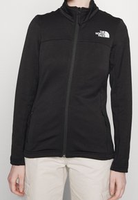 Czarna polarowa kurtka z wysokim kołnierzem, zamek błyskawiczny z przodu i kieszeniami po bokach. Posiada białe logo w górnej lewej części klatki piersiowej. Gładka faktura.