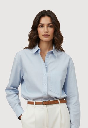 Vrouw met lang golvend haar, die een lichtblauw overhemd met knopen draagt, ingestopt in een witte broek met een bruine riem, staand tegen een effen achtergrond.