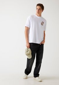 Camiseta blanca de algodón con logo en el pecho, combinada con jeans oscuros. El modelo sostiene una gorra verde clara y lleva zapatillas blancas. Look simple y casual.