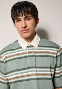 Polo majica svijetlo zelene boje s horizontalnim prugama u bijeloj, narančastoj i tamnosivoj boji. Ima kontrastni vratni ovratnik u boji kreme i prednju ploču s gumbima.