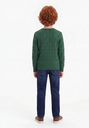 Bambino con capelli rossi e ricci visto di spalle, indossa un maglione verde a trecce, jeans blu e scarpe marroni su sfondo bianco.