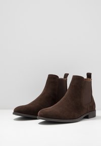 Botines de ante marrón con paneles laterales elásticos, suelas planas y tiradores. Textura suave y diseño de punta redondeada.