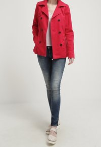 Chaqueta roja de doble botonadura con grandes botones negros. Tela lisa, cuello con muesca, combinada con jeans ajustados azules y zapatos beige.