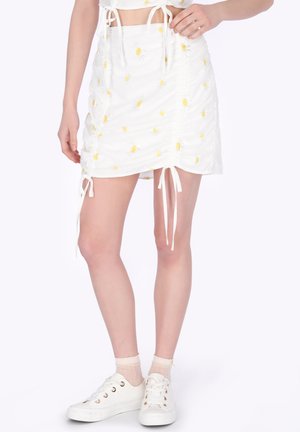 Biała spódnica z żółtymi nadrukami stokrotek i regulowanymi wiązaniami po bokach, noszona z białymi trampkami i przezroczystymi skarpetkami do kostek.