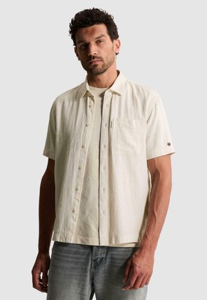 Homme aux cheveux courts et bouclés portant une chemise écrue à manches courtes boutonnée par-dessus un t-shirt assorti et un jean bleu clair, debout de manière neutre.
