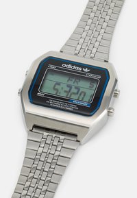 Montre numérique avec un bracelet en acier inoxydable, boîtier rectangulaire argenté, lunette noire et bleue, et un affichage numérique vert.