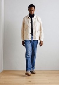 Veste beige légère avec de grandes poches, fermeture à boutons et texture douce, portée sur un col roulé noir, associée à un jean bleu et des chaussures marron.