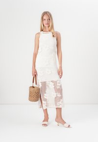 Bela cvetna pletena obleka z oprijetim korpusom in prosojnim, vzorčastim krilom. Povezana z tkanim bež nahrbtnikom in svetlimi petami.