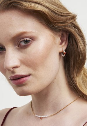 Kuldne kaelakee valgete helmeste ja punase aktsendiga; rõngaskõrvarõngad, millel on pärlid ja väike südamekujuline ripats.