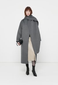 Grijze wollen jas met een brede kraag en diepe voorkant, gedragen over een crèmekleurige rok. Model houdt een klein zwart accessoire vast, met zwartehakkenlaarzen.