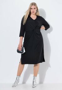 Vestido negro hasta la rodilla con detalle frontal retorcido, mangas tres cuartos y aberturas laterales. Combinado con botas tobilleras plateadas y un bolso metálico.