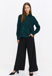 Czarna bluzka z długim rękawem w ciemnym odcieniu zieleni z wzorem spiralnym, w zestawieniu z szerokimi czarnymi spodniami i czarnymi szpilkami. 