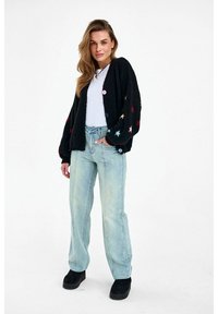 Woman stands wearing black cardigan with colored star patterns, white shirt, light blue jeans, and black platform shoes, hands in pockets.