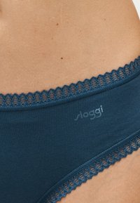 Gros plan sur la hanche d'une personne portant des sous-vêtements en coton bleu avec bordure en dentelle et un logo "sloggi" discret sur le tissu.
