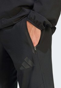 Čierne športové nohavice s vreckom na zips, hladkou textúrou a veľkým matným čiernym logom s tromi pruhmi na boku.