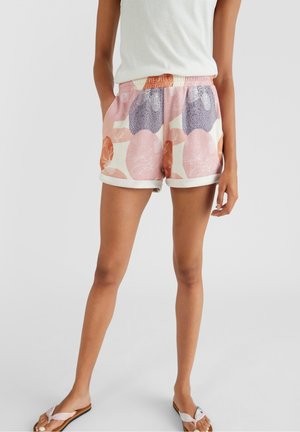 Kvinde iført pastelfarvede blomstrede shorts med hvide omsvøbte kanter, hvid top og hvide sandaler, stående foran en ensfarvet baggrund.