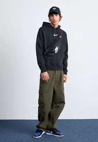 Musta huppari turkoosilla Nike-logolla, oliivinvihreät cargo-housut ja musta lippalakki. Musta crossbody laukku valkoisella Nike-logolla. Sinivalkoiset lenkkitossut.